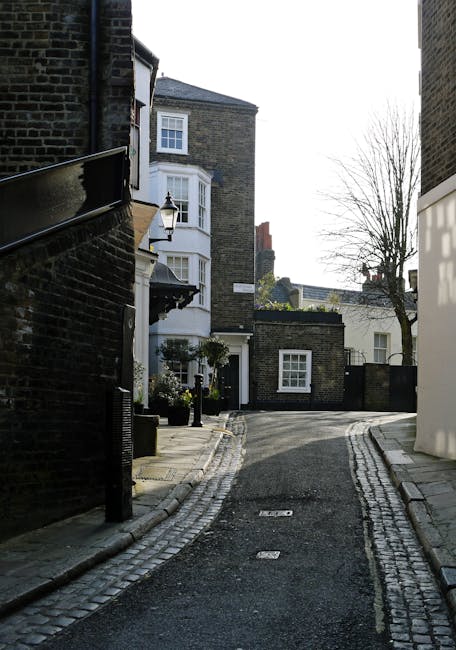 A narrow residential street in Hampstead with cobblestone paving and a gentle curve, lined with brick and white-painted houses featuring bay windows, some with small front gardens or potted plants. On