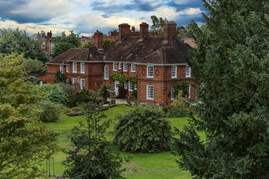 A large, historic brick building with multiple chimneys and a steeply pitched roof, surrounded by dense greenery including trees, shrubs, and a well-maintained lawn. The house features several white-framed windows and a small entrance garden with a wooden gate. The scene appears to be taken during daytime with partly cloudy skies overhead. In the context of house removals and transport, this image illustrates an exterior view of a residential property in Hampstead, often relevant to home relocation services offered by companies like Removal Companies Hampstead, highlighting the setting where furniture transport and packing processes may take place before a house move.