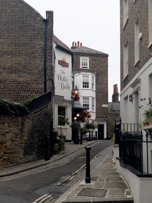 Close-up view of a brick wall displaying a rectangular metal street sign with the text 'LOWER TERRACE NW3' in white lettering on a dark green background. The sign is mounted to the wall, which is composed of red and brown bricks with visible mortar. In the background, slightly blurred, there is a street scene with greenery and trees, indicating an outdoor setting near a residential area in Hampstead. This scene is captured as part of a house removal or home relocation process, with the focus on exterior details such as signage and building materials. The environment appears to be well-lit, possibly during daylight hours, aligning with a professional moving service provider's documentation, like Removal Companies Hampstead, preparing for furniture transport and packing for a house move.
