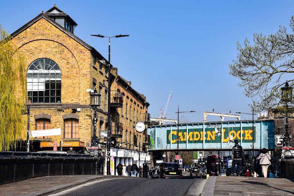 The image depicts a street scene in Hampstead, featuring a brick building with large arched and rectangular windows, outdoor street lighting, and a clock mounted on a black pole. Vehicles are parked a