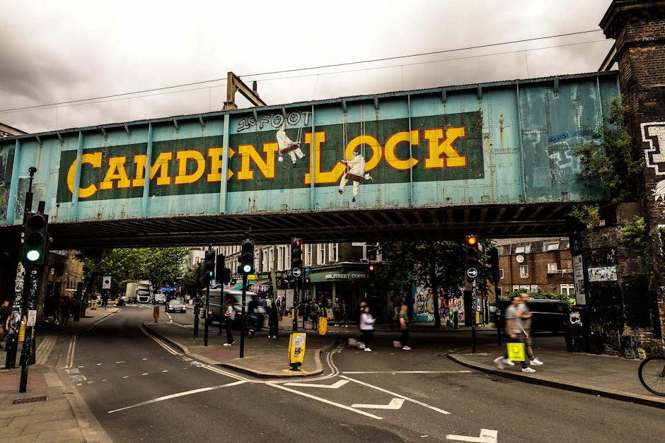 The image depicts a street scene in Hampstead, featuring a brick building with large arched and rectangular windows, outdoor street lighting, and a clock mounted on a black pole. Vehicles are parked a
