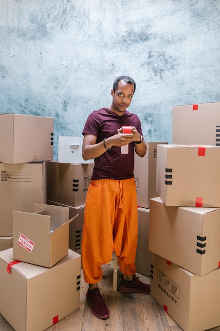 A man in casual clothing, wearing a maroon T-shirt and bright orange pants, stands amidst a collection of cardboard boxes in an indoor space with a textured, light blue wall background. He is holding a smartphone in both hands, appearing to be checking or communicating something. The boxes around him vary in size, some sealed with red or black tape, others open, revealing packing materials such as bubble wrap and plastic wraps. The scene captures a moment of packing or planning during a home relocation or moving process, with the man engaged in coordination or communication related to furniture transport and moving logistics, as part of a professional removal service. The environment suggests an organized packing area prepared for loading onto a moving vehicle, supporting the process of house removals.