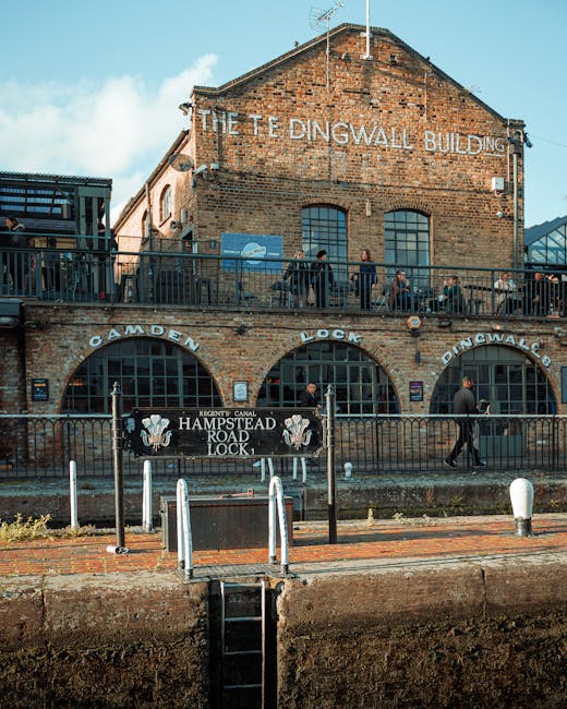 An exterior view of the historic Hampstead Road Lock building in Camden, featuring its brick facade and large arched windows. The building has a sign that reads 'The Tedingwall Building' on the upper section. A black metal railing surrounds the front area, with several people walking along the pavement and standing near the entrance. In front of the building, there is a sign for the 'Hampstead Road Lock' and a canal lock gate system with black gates, white bollards, and a small ladder leading down to the canal. The scene is set during daylight, with partly cloudy skies, and the setting suggests activity related to house removals or moving logistics, as part of a professional relocation service process. Removal Companies Hampstead could assist with relocating furniture and belongings in such urban environments. The image captures a moment of movement and preparation typical of home relocation procedures involving loading or unloading near historic waterways.