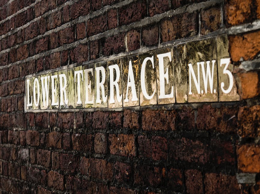 Close-up photograph of a street name plaque mounted on a red brick wall, reading 'Lower Terrace NW3' in white capital letters on a yellow background. The plaque appears weathered, with some dirt and moss visible around its edges. The brick wall behind it shows a pattern of irregular reddish-brown bricks with mortar in between, typical of older residential buildings in Hampstead. This image reflects the setting where house removals or packing and moving services may be required, providing contextual familiarity for clients seeking professional removal companies in Hampstead. The overall scene suggests a narrow or residential street, pertinent to planning home relocation or furniture transport in areas with narrow access, supporting the services offered by Removal Companies Hampstead.