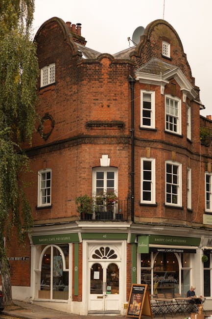 An exterior view of the historic Hampstead Road Lock building in Camden, featuring its brick facade and large arched windows. The building has a sign that reads 'The Tedingwall Building' on the upper section. A black metal railing surrounds the front area, with several people walking along the pavement and standing near the entrance. In front of the building, there is a sign for the 'Hampstead Road Lock' and a canal lock gate system with black gates, white bollards, and a small ladder leading down to the canal. The scene is set during daylight, with partly cloudy skies, and the setting suggests activity related to house removals or moving logistics, as part of a professional relocation service process. Removal Companies Hampstead could assist with relocating furniture and belongings in such urban environments. The image captures a moment of movement and preparation typical of home relocation procedures involving loading or unloading near historic waterways.