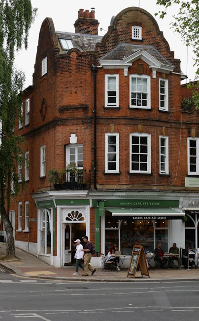 The image depicts a three-storey red-brick building located on a street corner, with a ground-floor bakery, cafe, and patisserie featuring a green awning. The upper floors display white-framed sash windows, some with small decorative balconies, and a rounded gable roof with chimneys and skylights. In front of the building, on the pavement, two men are engaged in carrying a large cardboard box, possibly part of a home relocation or moving process. Nearby, several pedestrians are walking along the sidewalk, while a few sit outside the cafe. A black trolley and some plastic-wrapped packages are visible near the entrance, indicating ongoing packing or furniture transport activities. The scene is set during daylight, with leafy trees framing the building and providing a sense of neighbourhood ambiance. This image is relevant to house removals and moving services offered by Removal Companies Hampstead, illustrating urban logistics and packing procedures involved in residential moves.