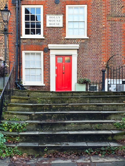 The image shows the front entrance of a traditional brick building labeled 'Holly Mount House' with a white sign attached to the upper wall. The building features a white-framed window above the door, and two additional white-framed windows located on either side of the entrance, all with closed blinds. The entrance has a vibrant red wooden door with a narrow vertical window and a letterbox, framed by a white lintel and surround. In front of the building, there are stone steps leading up to the door, with some moss and small plants growing between the treads. To the right of the doorway, there is a small potted shrub in a green container, and a black metal railing runs along the sides of the steps, providing support and safety. This scene captures the exterior of a residential property that could be involved in a house removal or home relocation process, with the surrounding environment featuring the typical urban pavement and outdoor fixtures.