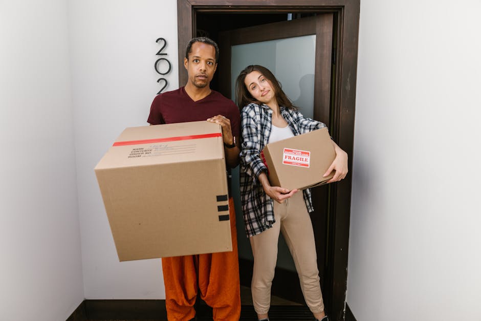 Two individuals, a man and a woman, are positioned inside a doorway of a residential property, preparing for a home relocation process. The man, on the left, has short dark hair and is dressed in an orange outfit, holding a large cardboard box sealed with red tape. The woman, on the right, has long brown hair and is wearing a plaid shirt over a light top, carrying a smaller cardboard box labeled 'Fragile' with red and white tape. Both are standing on a black flooring surface in a narrow hallway or entrance area. The background shows a closed door frame with the house number partially visible on the wall beside it, and the environment appears well-lit with indoor lighting. This image, associated with Removal Companies Hampstead, visually depicts packing and loading activities typical of professional removals, highlighting packaging materials, furniture transport, and the careful handling of fragile items during a house move.